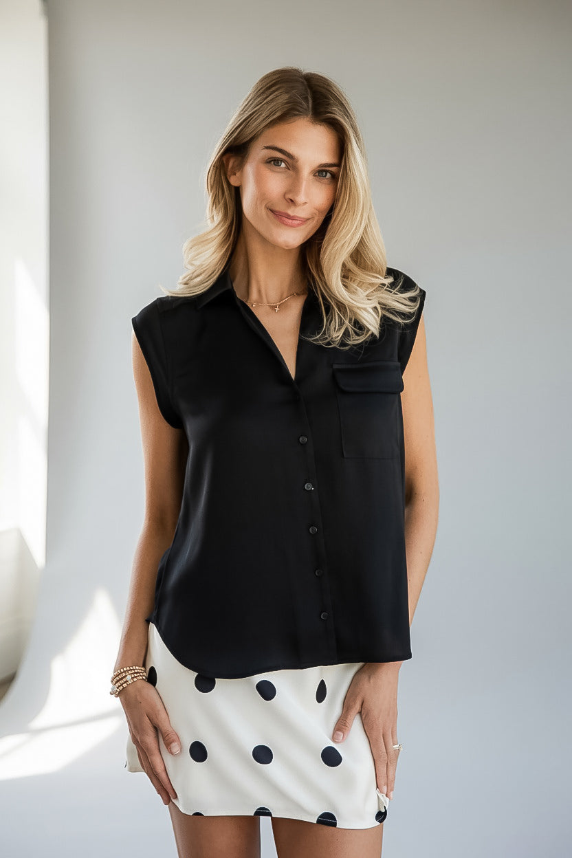 A woman modeling a sleeveless black button-front blouse with a collar and chest pocket, paired with a white skirt featuring black polka dots, set against a neutral light gray background.