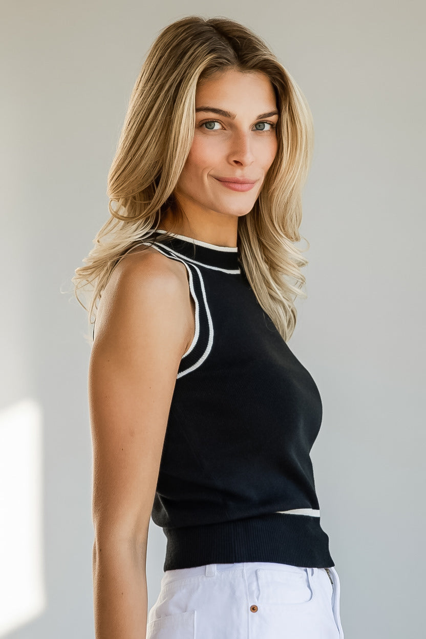 A woman wearing a sleeveless black knit top with white contrast trim and white pants, photographed against a plain light gray background.