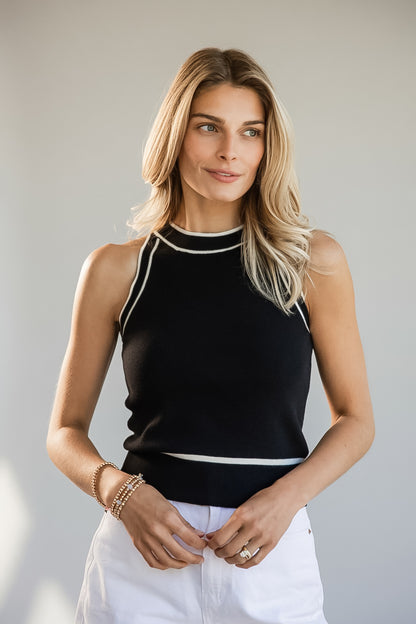 A blonde woman wearing a sleeveless black knit top with white contrast trim and white pants, standing against a plain light gray studio background.
