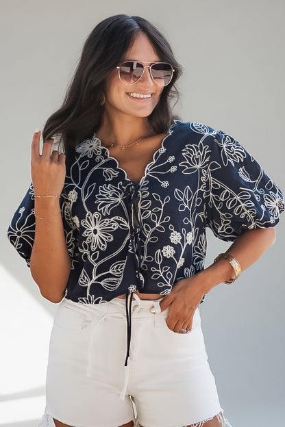 A woman wearing a navy blue short-sleeve blouse with a white floral line-art pattern, paired with white shorts, photographed against a plain light gray background.