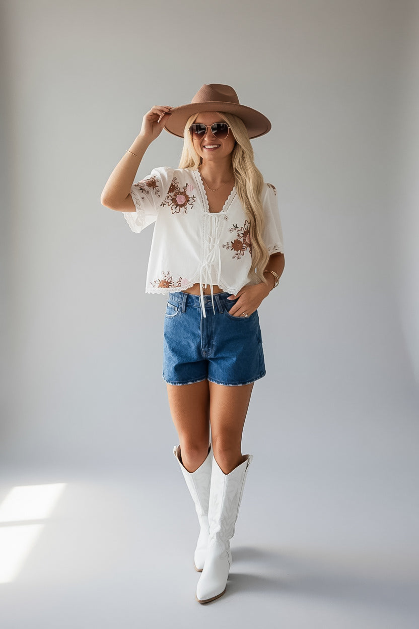A woman modeling a bohemian-style outfit consisting of a white short-sleeve embroidered blouse, blue denim shorts, white cowboy boots, a brown wide-brim hat, and sunglasses, set against a plain light gray studio background.