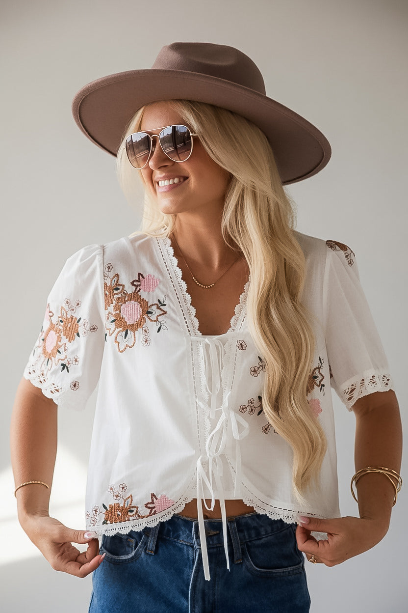 A woman wearing a white short-sleeve bohemian crop top with floral embroidery and lace trim, paired with a brown wide-brim hat and blue jeans.