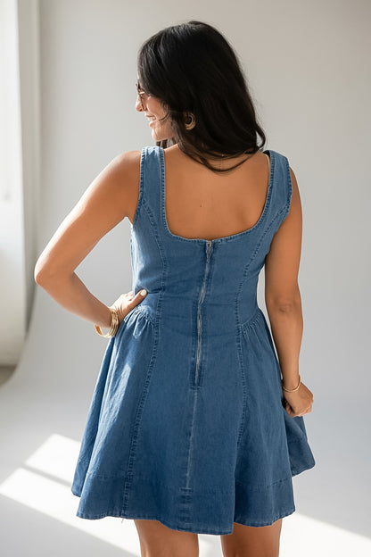 A woman wearing a blue denim sleeveless dress, photographed from behind to show the back zipper and fit, standing in a bright indoor setting.