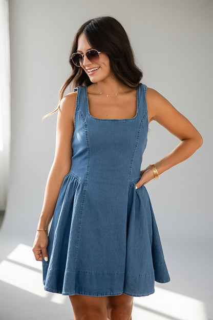A woman modeling a sleeveless blue denim dress with a square neckline and a flared skirt, standing in a bright indoor setting with sunlight on the left.