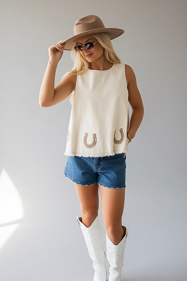 A woman modeling a sleeveless cream top with embroidered horseshoes, paired with denim shorts, white cowboy boots, and a tan wide-brim hat against a light gray studio background.