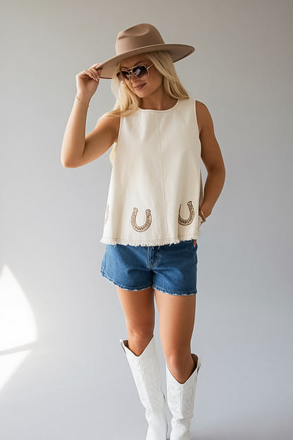 A woman modeling a sleeveless cream top with embroidered horseshoes, paired with denim shorts, white cowboy boots, and a tan wide-brim hat against a light gray studio background.