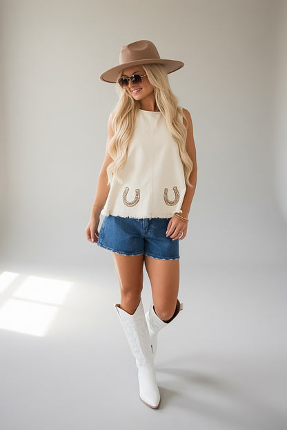 A woman modeling a sleeveless cream top with two horseshoe details, paired with denim shorts, white cowboy boots, and a brown wide-brim hat against a light gray studio background.