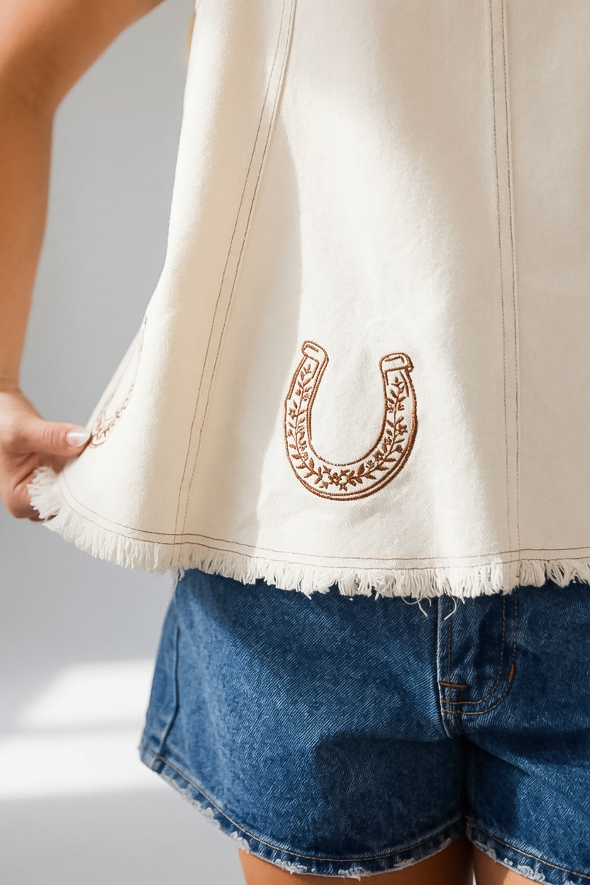 Close-up of a person wearing a cream-colored sleeveless top with a brown embroidered horseshoe design on the front, paired with blue denim shorts.