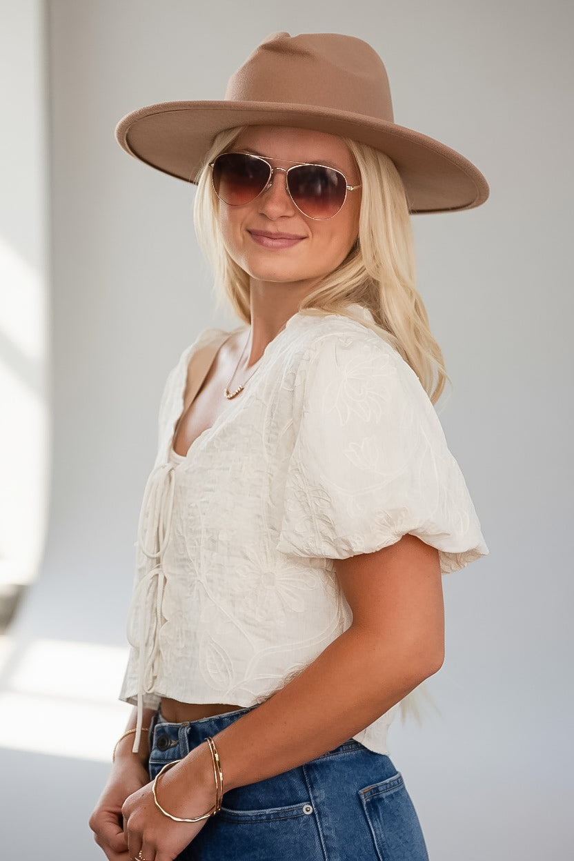 A woman wearing a cream-colored textured puff-sleeve crop top, blue jeans, a tan wide-brim fedora hat, and sunglasses, posing in a bright indoor setting.