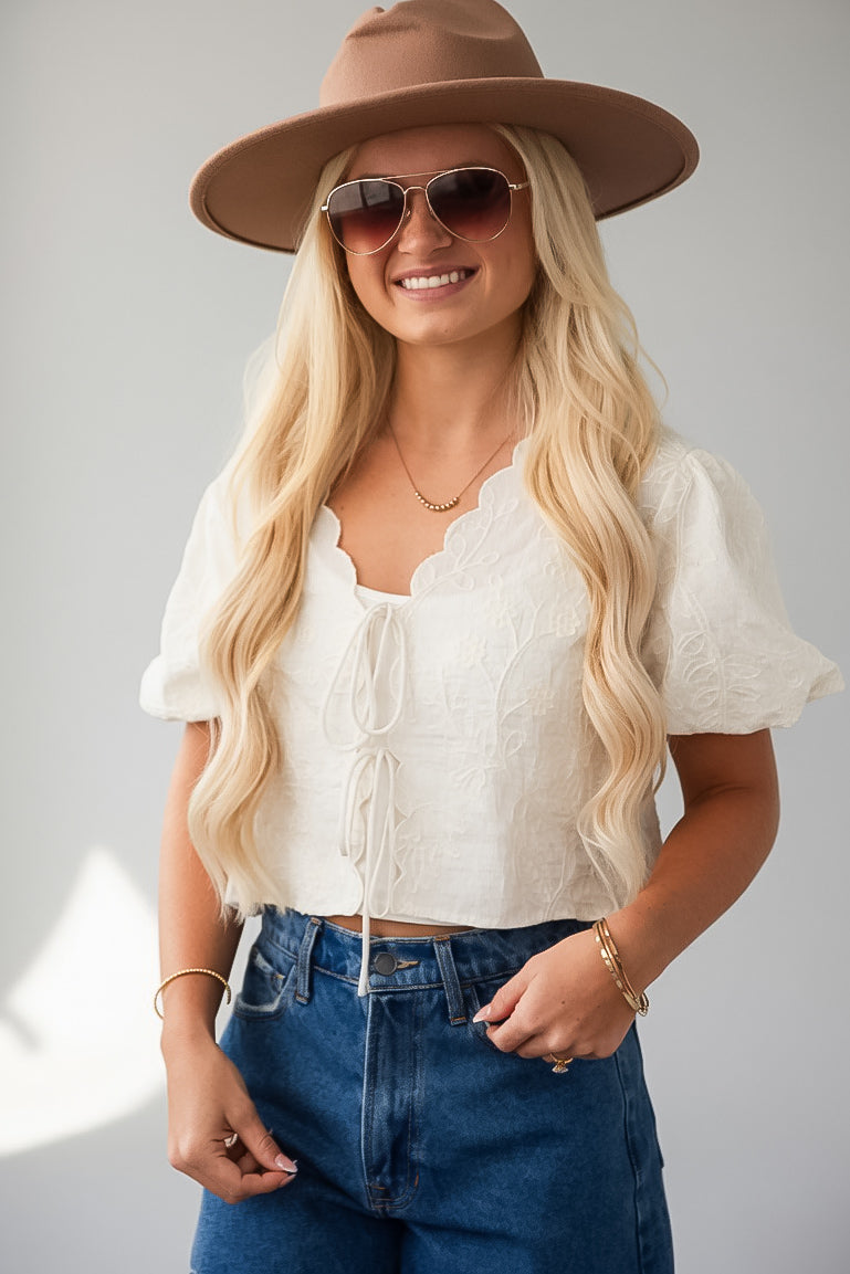 A woman wearing a cream lace short-sleeve crop top, blue jeans, a brown wide-brim hat, and sunglasses, posing against a light gray background.