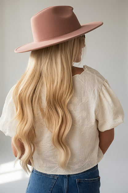 A woman with long blonde wavy hair is shown from behind, wearing a dusty pink wide-brim fedora hat, a cream textured short-sleeve blouse, and blue jeans against a light gray background.