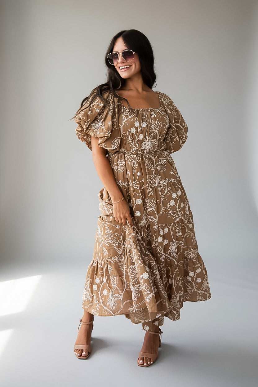 A woman modeling a brown bohemian-style midi dress with a white floral vine pattern, puff sleeves, and an off-the-shoulder neckline, paired with brown strappy high-heeled sandals against a neutral gray studio background.