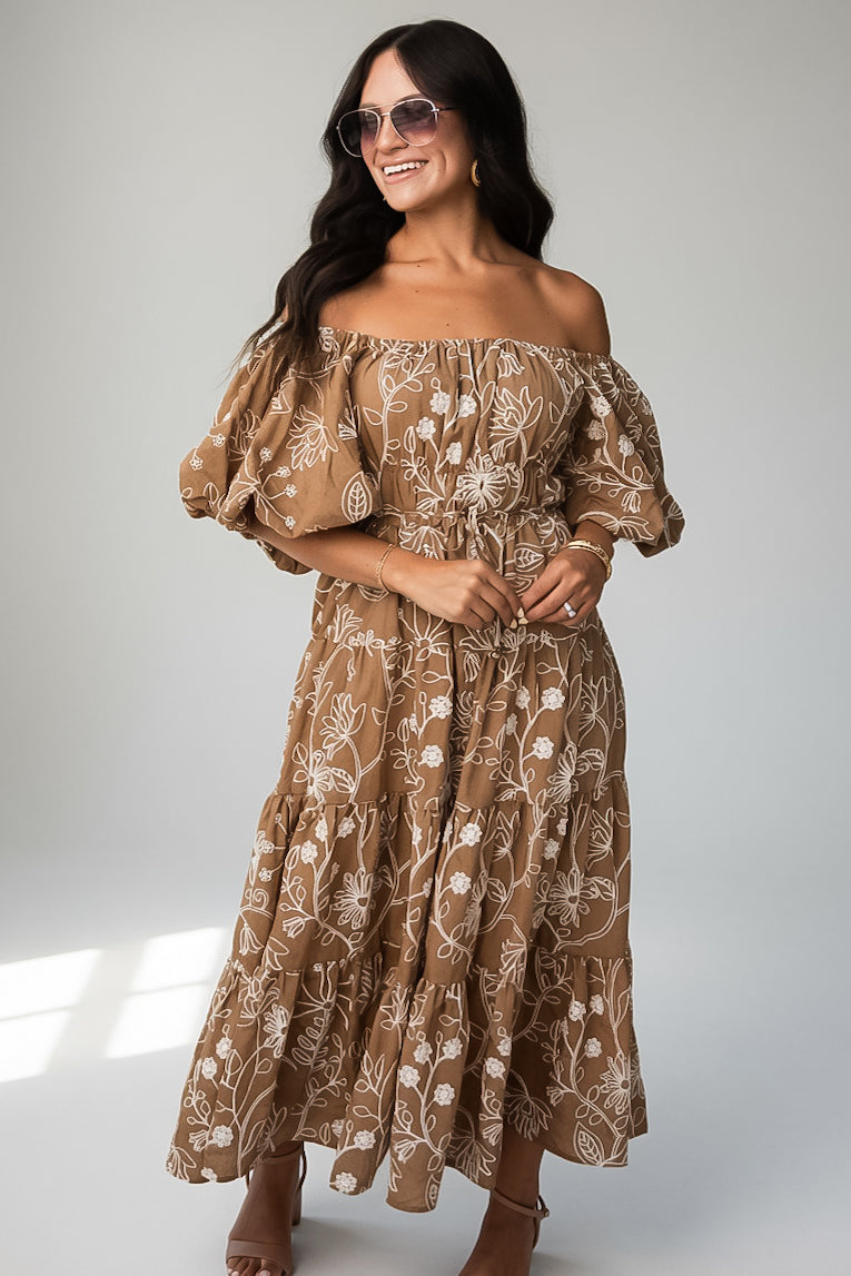 A woman modeling an off-the-shoulder brown maxi dress with a white floral pattern, styled with sunglasses and neutral heels against a light gray studio background.