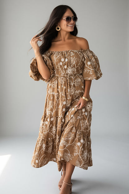 A woman modeling an off-the-shoulder midi dress with puff sleeves and an allover white floral print on a warm tan background, photographed against a light gray studio backdrop.