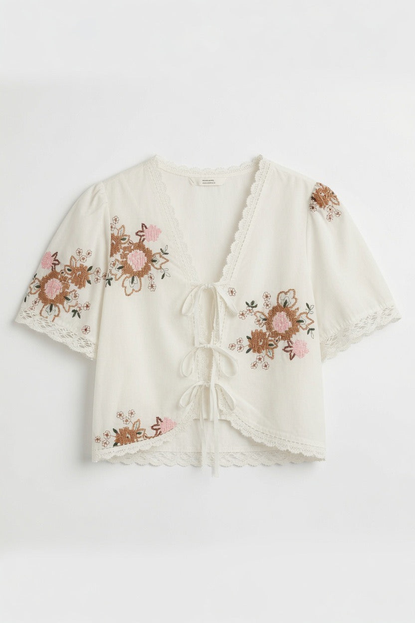 An off-white short-sleeve crop top with pink and brown floral embroidery, lace trim, and a front tie closure, shown flat on a white background.