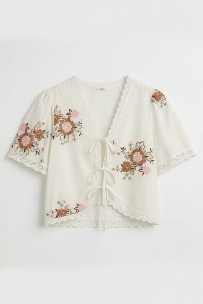 An off-white short-sleeve crop top with pink and brown floral embroidery, lace trim, and a front tie closure, shown flat on a white background.