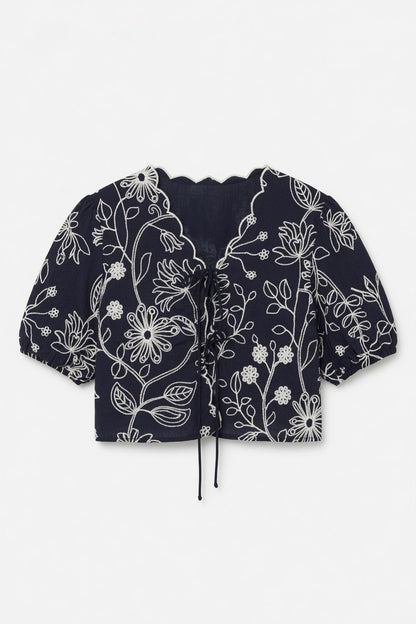 A cropped short-sleeve blouse in navy blue with a white floral line-art pattern, featuring a ruffled V-neckline and a drawstring tie at the front, laid flat against a light gray background.