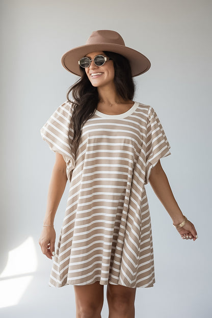 A woman wearing a beige and white horizontally striped short-sleeve tunic dress, a brown wide-brim hat, and round sunglasses, standing against a light gray studio background.