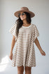 A woman wearing a beige and white horizontally striped short-sleeve tunic dress, a brown wide-brim hat, and round sunglasses, standing against a light gray studio background.