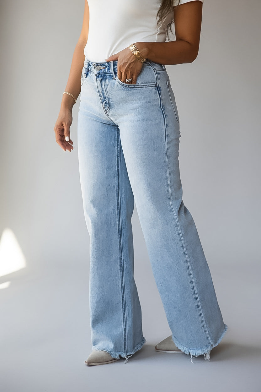 A person wearing light-wash wide-leg denim jeans with a frayed hem, paired with a white top and pointed shoes, photographed against a plain light gray studio background.