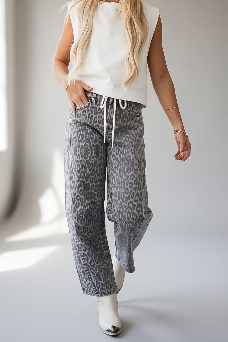 A woman wearing gray leopard-print barrel-leg pants with a white drawstring waist, paired with a white sleeveless top and white pointed-toe boots, shown in a bright indoor setting.