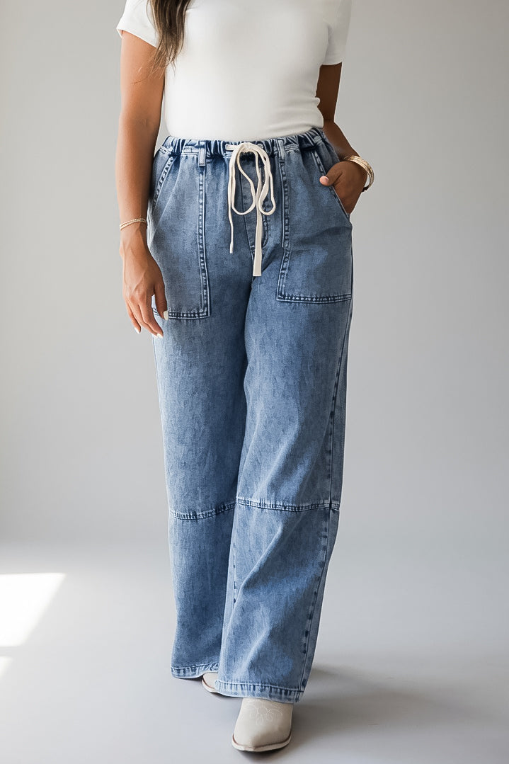 A person wearing light blue wide-leg denim jeans with a drawstring waist, paired with a white top and white shoes, standing against a light gray studio background.