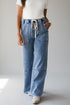 A person wearing light blue wide-leg denim jeans with a drawstring waist, paired with a white top and white shoes, standing against a light gray studio background.