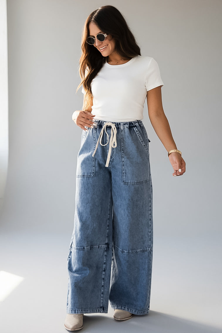 A woman modeling a pair of wide-leg blue denim jeans with a drawstring waist, paired with a white t-shirt and sunglasses, standing against a light gray studio background.