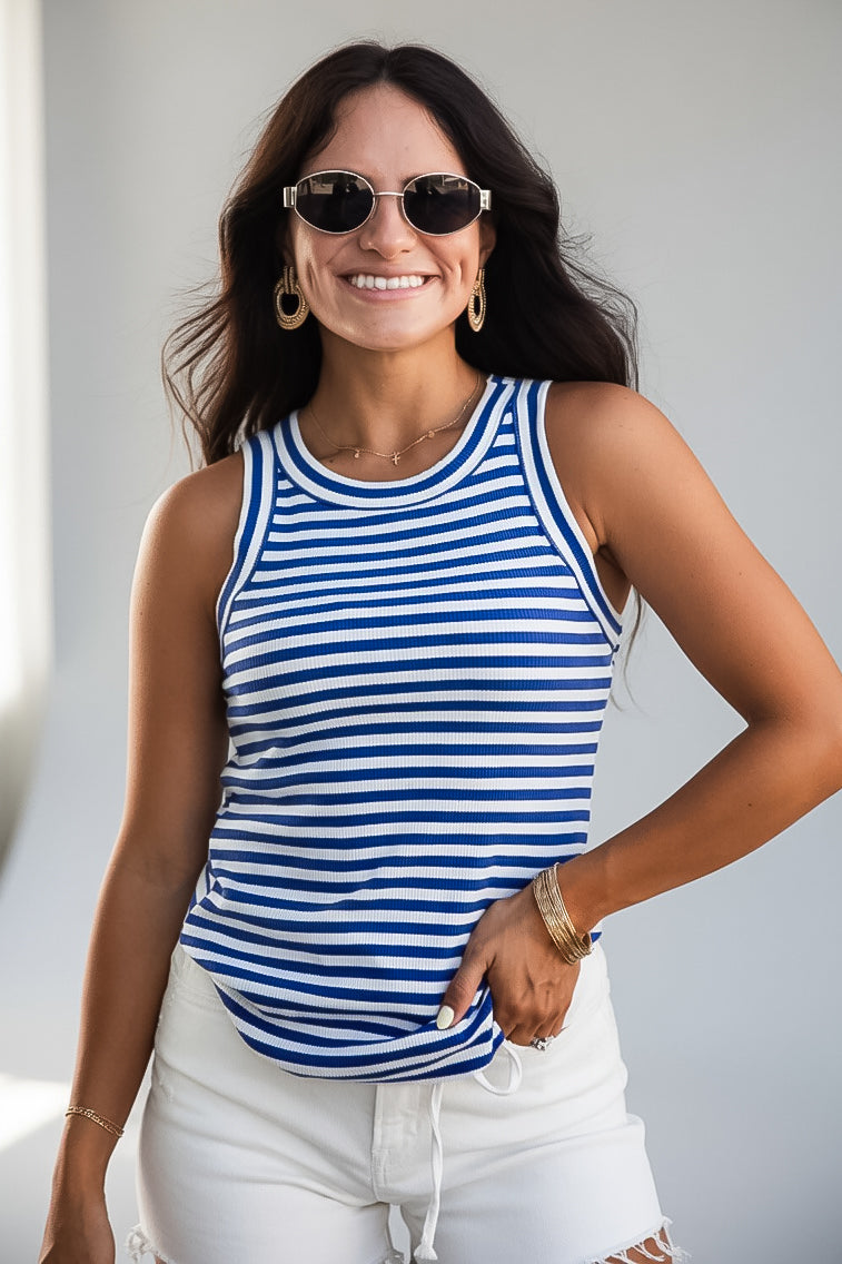 A woman wearing a blue and white horizontally striped tank top, white shorts, and sunglasses, posing with one hand in her pocket against a light gray background.