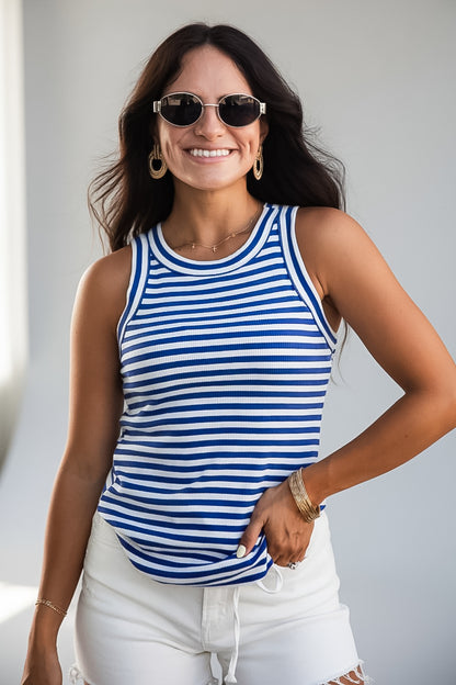 A woman wearing a blue and white horizontally striped tank top, white shorts, and sunglasses, posing with one hand in her pocket against a light gray background.