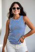 A woman wearing a blue and white horizontally striped tank top, white shorts, and sunglasses, posing with one hand in her pocket against a light gray background.