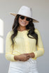A woman wearing a pale yellow ribbed long-sleeve button-front top, a white wide-brim fedora hat, and white pants, photographed against a light gray studio background.