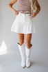 A model wearing a white denim ruffled mini skirt, a brown and white striped tank top, and white cowboy boots, standing against a light gray studio background.
