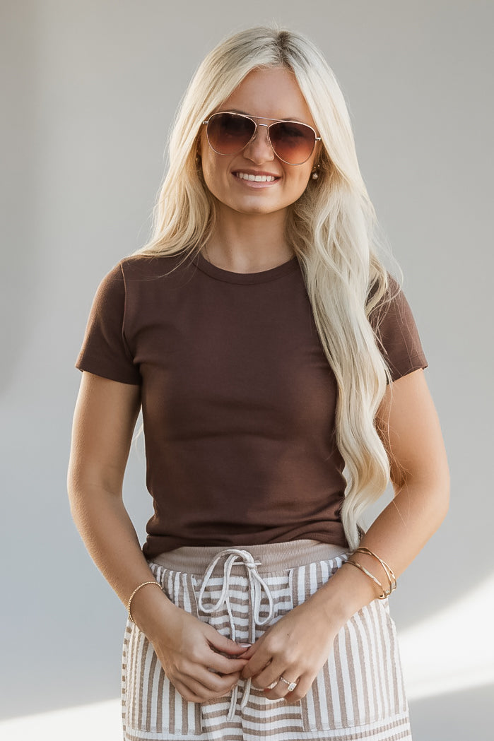 A woman with long blonde hair wearing a brown short-sleeve crewneck t-shirt and striped drawstring shorts, accessorized with sunglasses and gold bracelets, posing against a neutral light gray background.