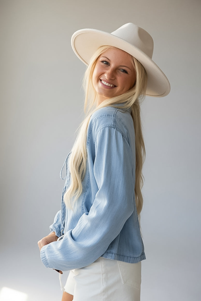 A smiling blonde woman wearing a wide-brimmed cream hat, a light blue denim jacket, and white pants, photographed against a plain light gray studio background.