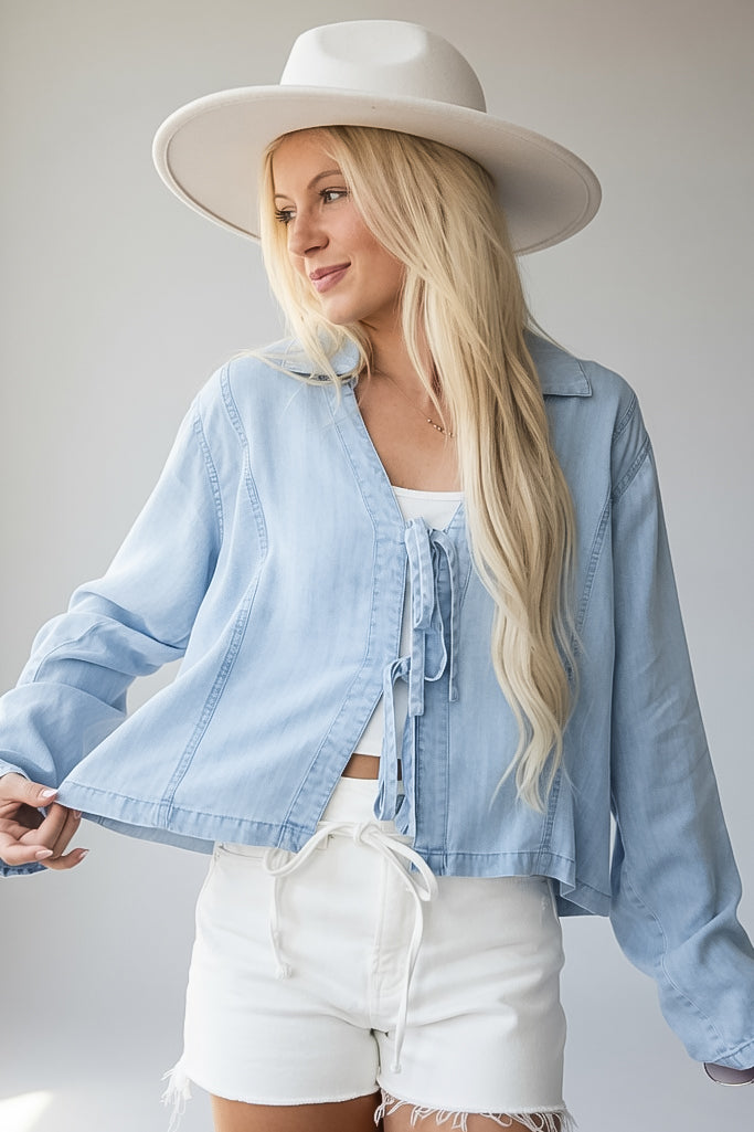 A woman wearing a light blue cropped denim jacket with front tie details, white shorts, and a wide-brim white hat, photographed against a plain light gray studio background.