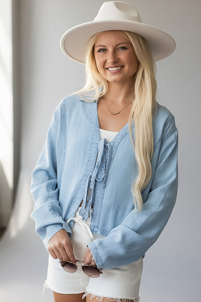 A smiling blonde woman wearing a light blue tie-front overshirt, white shorts, and a wide-brim white hat, holding sunglasses against a soft light gray indoor background.