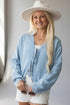 A smiling blonde woman wearing a light blue tie-front overshirt, white shorts, and a wide-brim white hat, holding sunglasses against a soft light gray indoor background.