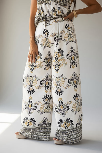 A model wearing a pair of wide-leg pants with a white base and an intricate black and gold damask-style floral pattern, paired with a matching patterned top.