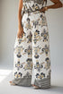 A model wearing a pair of wide-leg pants with a white base and an intricate black and gold damask-style floral pattern, paired with a matching patterned top.