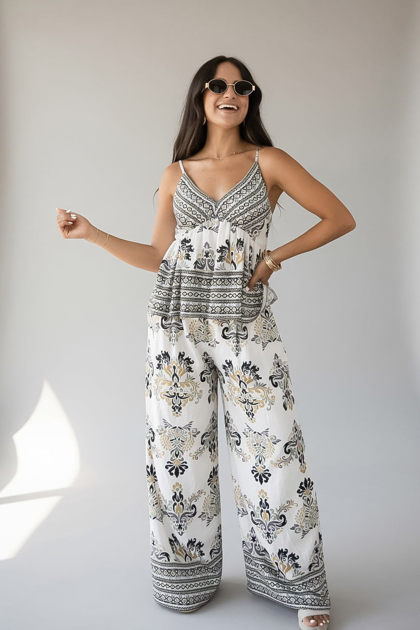 A woman modeling a sleeveless white jumpsuit with a black and beige bohemian-style floral and geometric pattern, set against a plain light gray studio background.