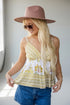 A woman wearing a bohemian-style white and yellow patterned camisole top, blue denim shorts, a dusty pink wide-brim hat, and sunglasses, standing in a brightly lit room.
