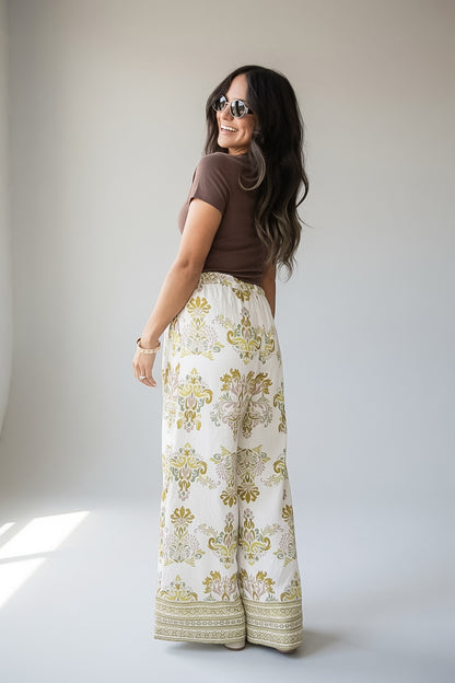 A woman wearing a brown short-sleeve top and high-waisted, wide-leg cream pants with a gold and green damask pattern, photographed in a studio setting.