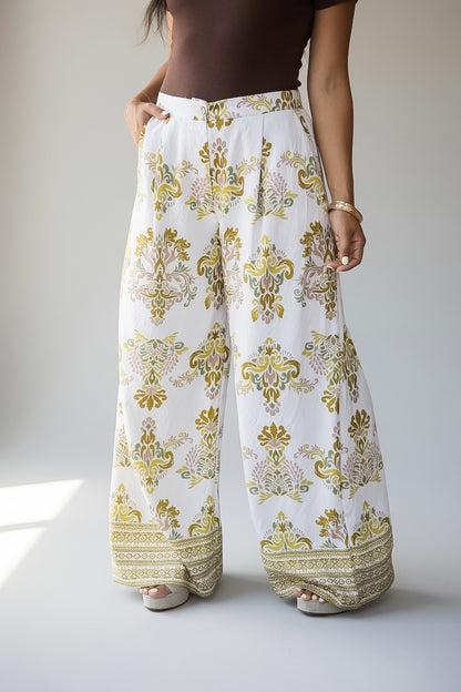 White wide-leg pants with an intricate gold and beige paisley pattern and decorative borders at the hems, shown on a model.