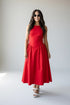 A woman modeling a sleeveless red midi dress with a fitted bodice and flared skirt, paired with white heeled sandals and sunglasses, standing against a plain light gray studio background.