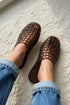 A person wearing brown woven slip-on mule shoes with rolled-up blue jeans, standing on a white textured rug.