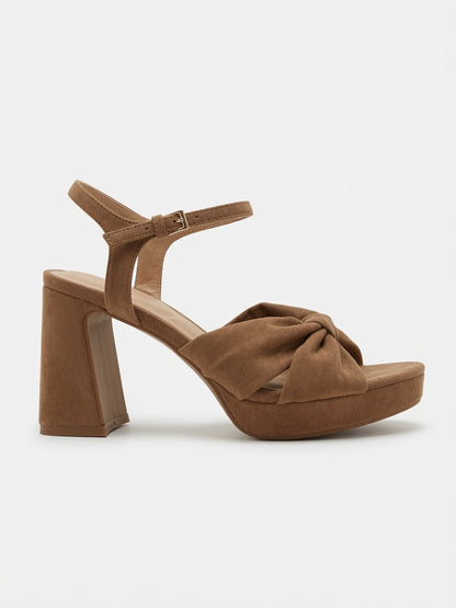 A single brown suede platform sandal with a chunky block heel, ankle strap, and a knotted bow detail on the front, shown against a plain light gray background.