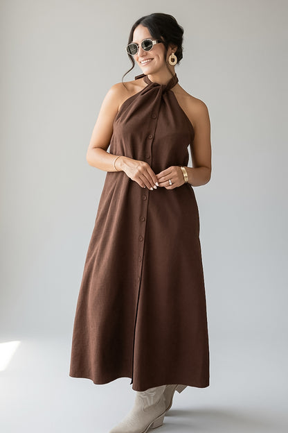 A woman modeling a brown halter-neck midi dress with a button-front closure, photographed against a plain light gray studio background.