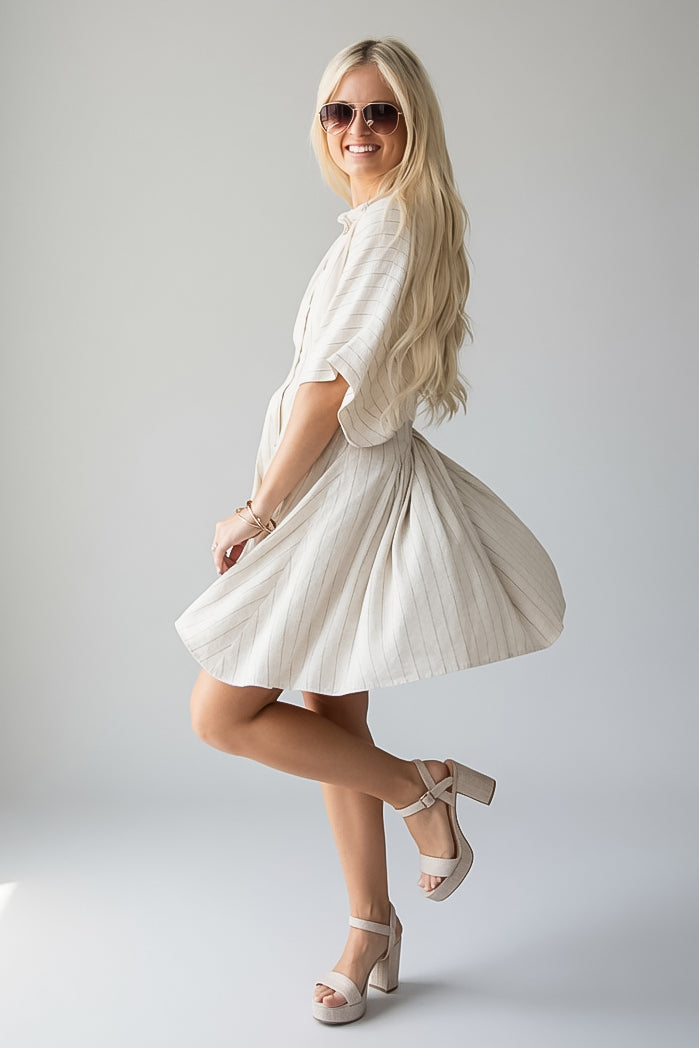 A woman modeling a short-sleeve cream linen dress with a flared skirt, paired with beige platform sandals and sunglasses, posing against a light gray studio background.
