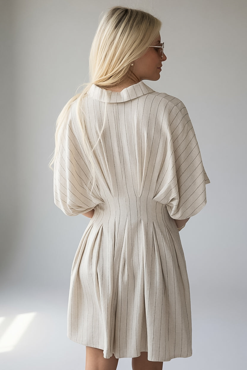 A woman with blonde hair is shown from the back wearing a pleated, short dress with wide three-quarter sleeves. The dress is a light cream or off-white color with thin vertical stripes, set against a plain light gray studio background.